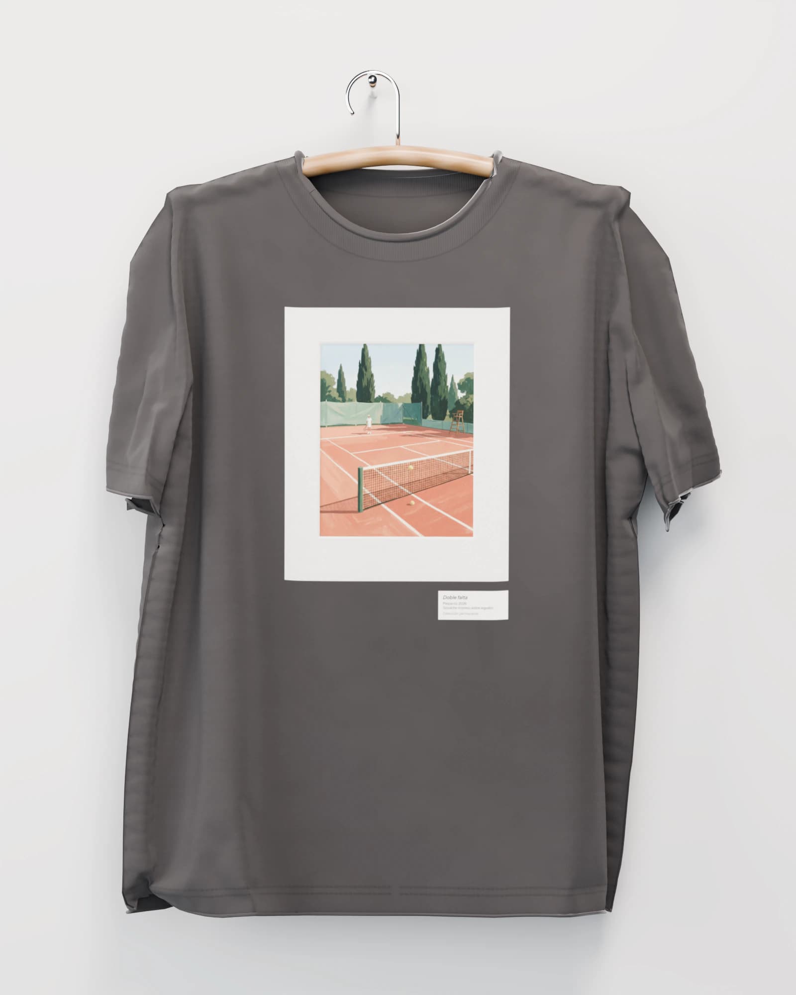 Polera color grafito lavado colgada de una percha de madera en un muro claro, con una cancha de arcilla con un tenista lejos y una pelota atrapada en la red, en marco negro con paspartú en el pecho