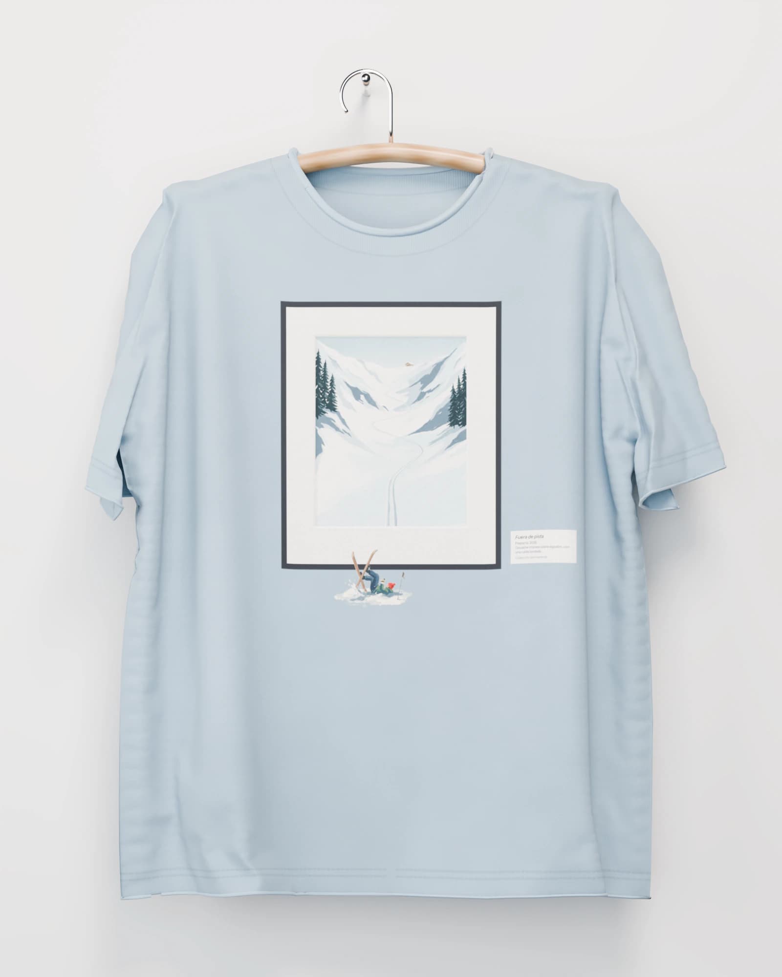 Polera color azul glaciar colgada de una percha de madera en un muro claro, con una ladera nevada con huellas de esquí, en marco negro, y un esquiador caído estampado bajo el marco en el pecho