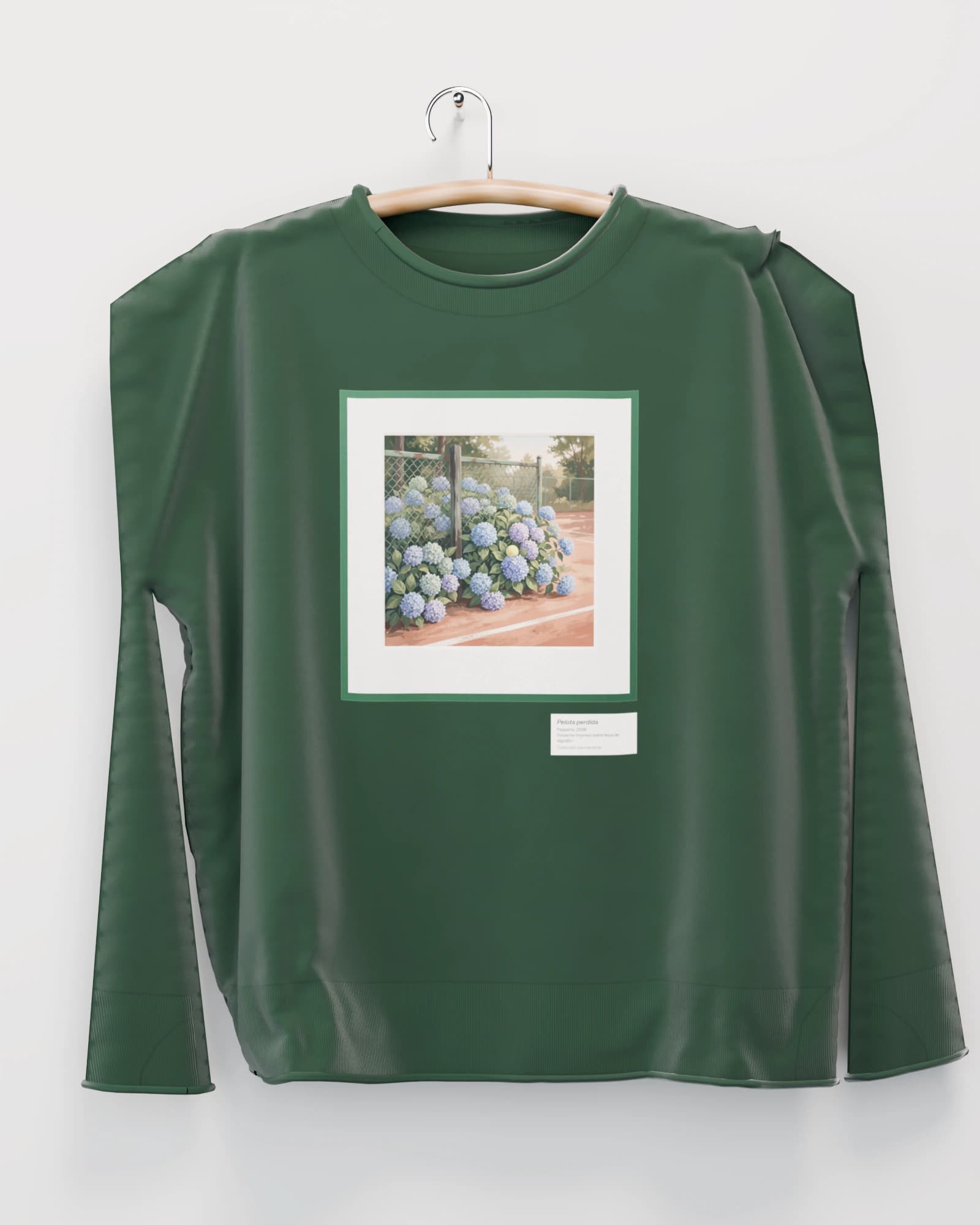 Polerón color verde cancha colgada de una percha de madera en un muro claro, con hortensias contra la reja de una cancha con una pelota escondida, en marco verde en el pecho