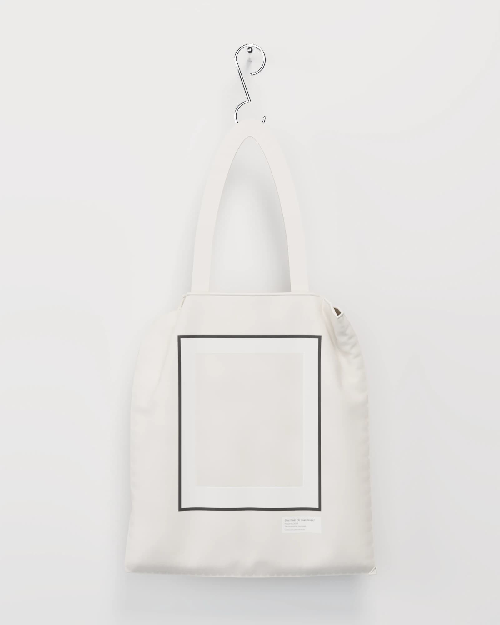 Bolso de lona cruda colgado de un gancho cromado por sus asas, con un marco negro vacío estampado y su cartela